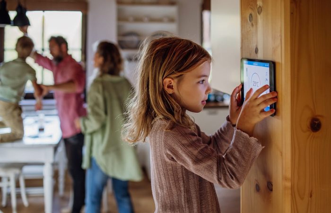 A young girl interacts with a smart home control panel mounted on a wall, while her family is seen in the blurred background of their home.